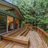 A green home has a spacious wooden deck that looks out to a wooded area.