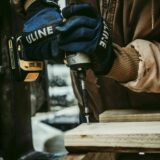 Close-up of a professional with protective blue gloves drilling a nail into a deck board.