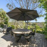 An outdoor dining setup on a sunny patio features a table surrounded by chairs and shaded by a large patio umbrella.